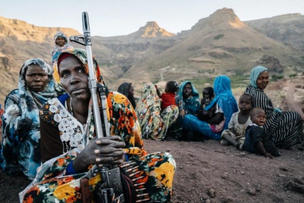 A group of women, some dressed in colorful traditional clothing, sit on rocky terrain in a mountainous area, with one woman prominently holding a rifle. The landscape behind them features steep hills under a clear sky.
