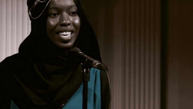 A young woman in a hijab stands at a microphone, smiling while speaking to an audience during a performance. The image is in black and white, emphasizing her expression and the stage lights behind her.