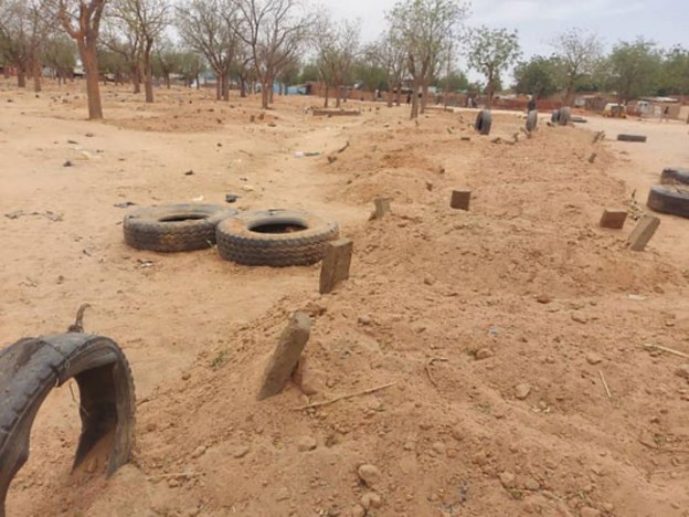 A barren landscape with scattered trees, featuring trenches and piles of dirt, alongside used tires, indicative of a desolate area, possibly related to conflict or abandonment.