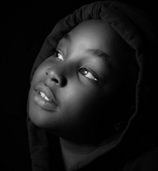 A close-up black and white portrait of a young girl looking upwards, wearing a dark hooded garment.