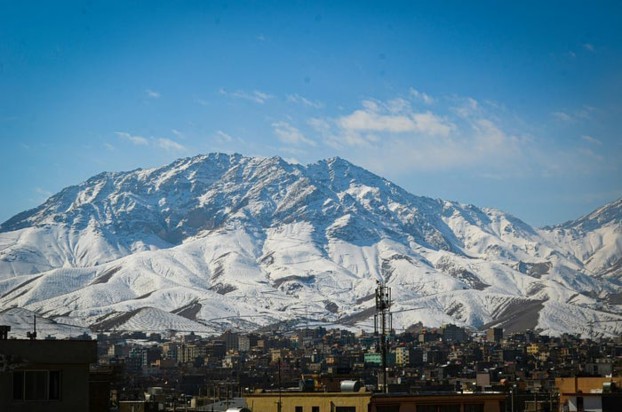 A panoramic view of a snowy mountain range against a clear blue sky, overlooking a city in Afghanistan.