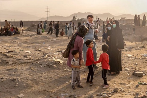 A group of people in a dusty environment with a backdrop of mountains. A woman dressed in traditional attire interacts with children, while adults stand nearby, some wearing masks. The setting appears to reflect a somber atmosphere, possibly in a context of displacement or hardship.