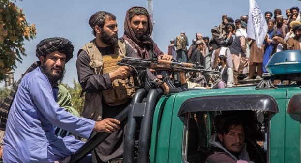 Taliban militants occupy the back of a military vehicle, displaying weapons and flags while surrounded by onlookers in a public area.