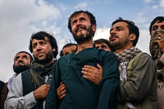 A group of Afghan men expressing deep emotion, with one man visibly distressed, surrounded by others who appear to offer support. The scene captures a moment of collective grief or remembrance against a cloudy sky.