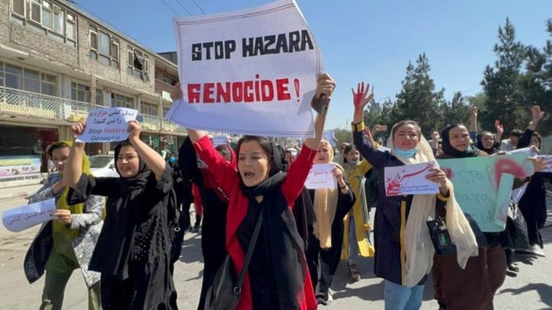A group of women in traditional attire march down a street holding signs that read 'STOP HAZARA GENOCIDE!' while raising their hands in protest.