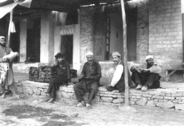 Historical black and white photograph depicting four Afghan men sitting on a stone wall in a rural setting, with traditional clothing and a rustic building in the background.