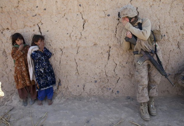 A soldier stands beside two young Afghan girls, all with their faces covered, against a cracked earth wall, depicting a tense and fearful moment amidst a backdrop of conflict.