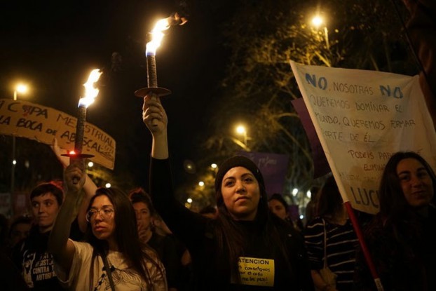 A group of women participating in a nighttime protest, holding lit torches and banners with messages advocating for women's rights and equality.