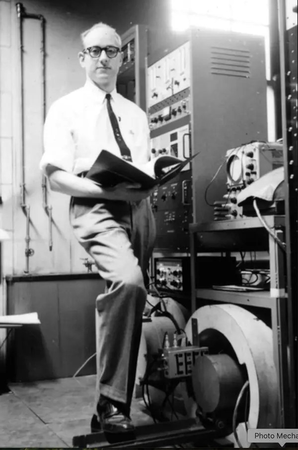 A man in glasses stands in a laboratory, holding an open book, with various scientific equipment and instruments visible in the background.