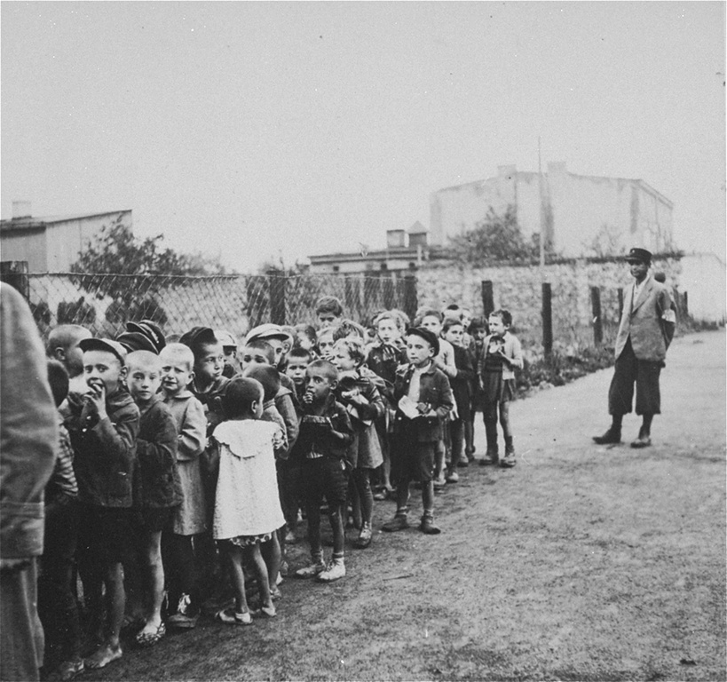 A black and white historical photograph depicting a line of children, some appearing malnourished, standing in front of a fence. A man in a suit watches from the side, as they await an uncertain fate in a bleak environment.