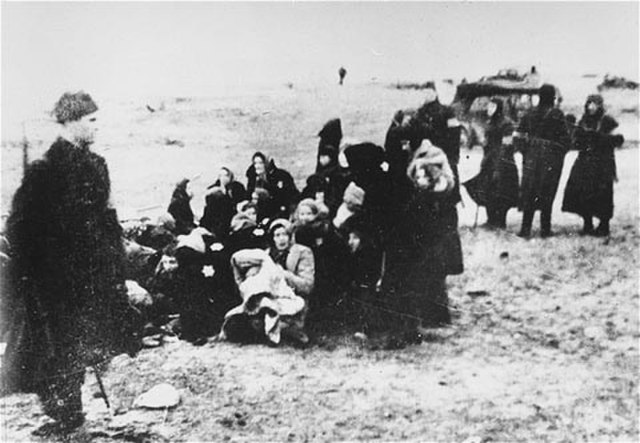 A group of Jewish women and children sit on the ground, appearing frightened and huddled together, as a German soldier stands guard nearby on a beach.