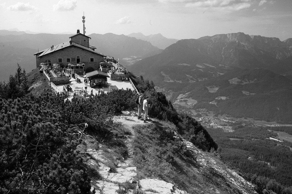 A scenic view of a mountain lodge perched on a rocky peak, surrounded by lush greenery and forest, with several people enjoying the outdoor seating area, set against a backdrop of majestic mountains under a clear sky.