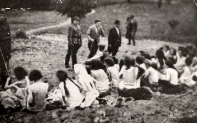 Black-and-white historical photograph of a group of women sitting on the ground, some dressed in light clothing, while men in military and civilian attire stand nearby overseeing the scene.