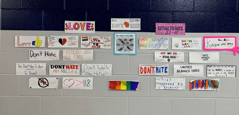 A colorful display of anti-hate messages and signs on a wall, featuring slogans like 'Love,' 'Don't Hate,' 'Haters Are Not Cool,' and 'United Against Hate,' along with drawings and symbols promoting love and respect.