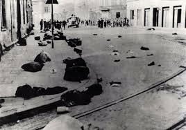 A historical black and white photograph showing a street littered with clothing and belongings, with several figures in the background gathered at a distance, illustrating a scene of aftermath and abandonment.
