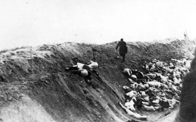 A historical black and white photograph showing a mass grave with bodies lying on the ground, while a solitary figure walks along the edge of the trench.