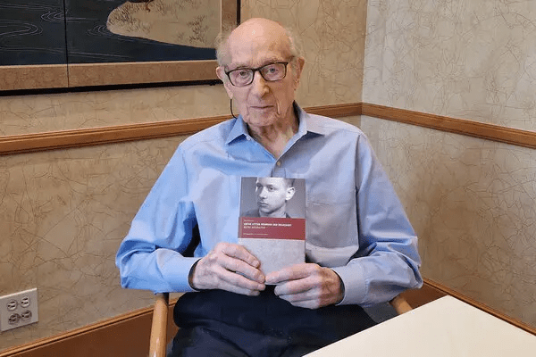 An elderly man with glasses holds a book titled 'The Last Survivor: Portrait of Edward Anders' while sitting at a table in a well-lit room.