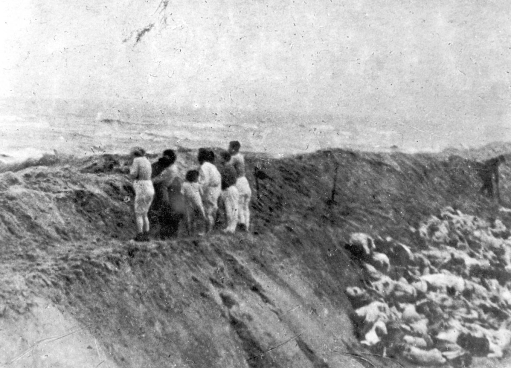 A group of individuals stands at the edge of a trench, viewed from behind, as they witness a stark scene of bodies lying in a mass grave nearby.