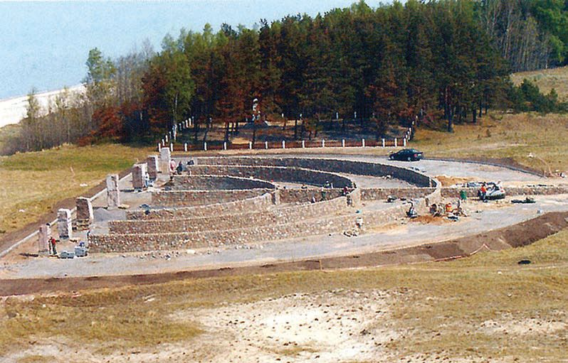 Aerial view of a memorial site under construction, featuring a circular stone structure surrounded by trees, located near a beach.