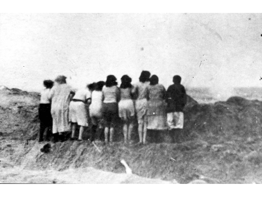 A black and white photograph depicting a group of women standing with their backs to the camera, observing a bleak landscape. They appear to be gathered at the edge of a trench, with sand dunes in the background, alluding to a somber historical context.