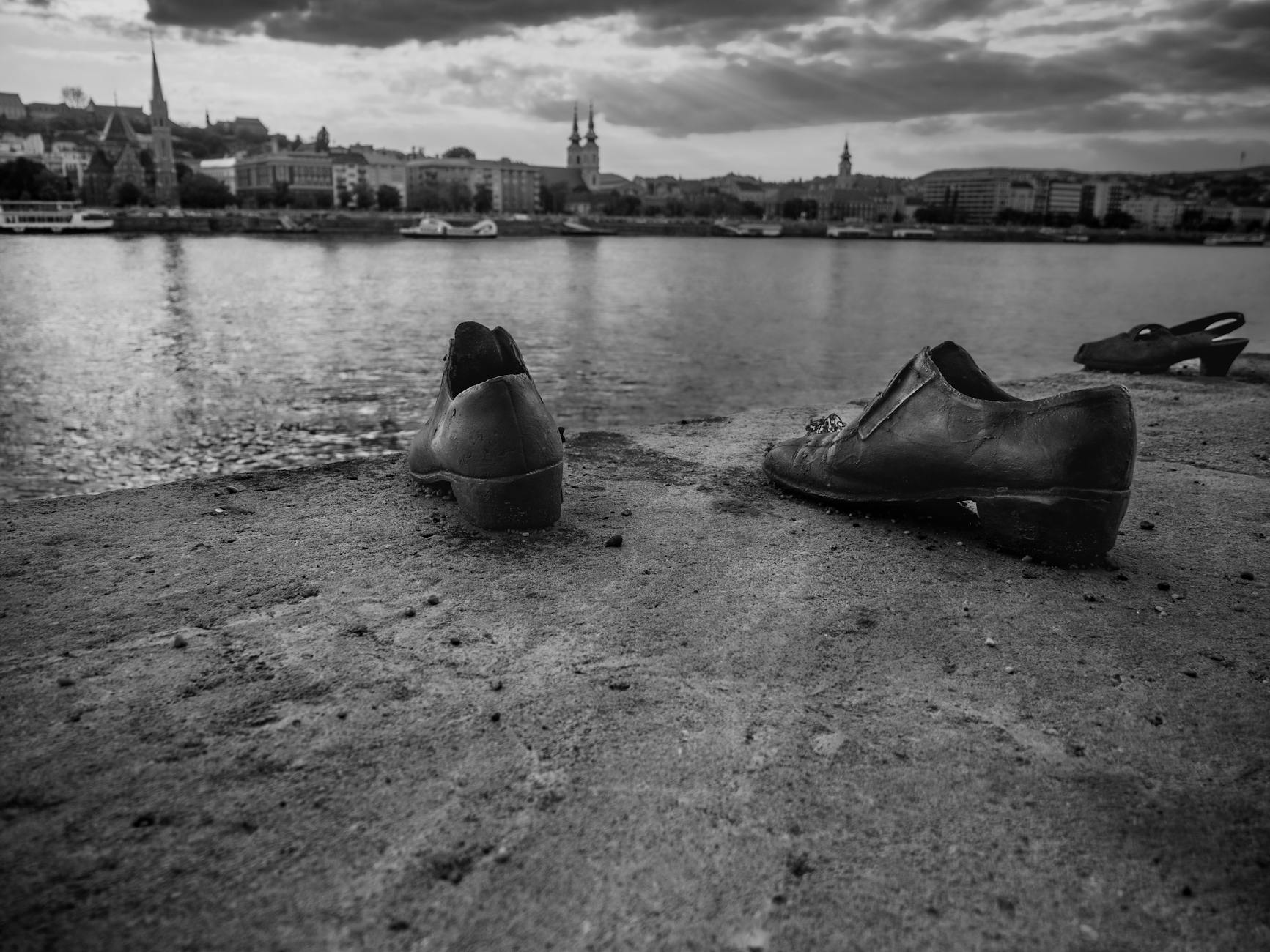 Black and white image of old shoes left on the ground near a river, with a city skyline in the background under a cloudy sky.
