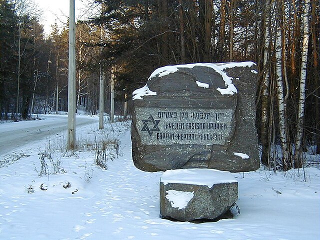 A stone memorial in a snowy landscape, inscribed in Hebrew and Yiddish, commemorating the victims of a massacre. The surrounding trees are bare, indicating winter, and a snow-covered road is visible in the background.