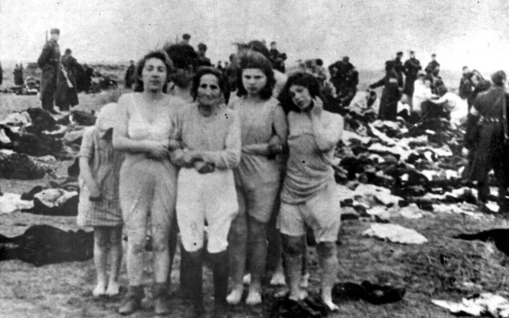 Five women standing in front of a mass of clothing, a grim scene indicating a historical atrocity, with armed soldiers visible in the background.